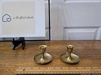 Pair of solid brass taper candle votives displayed side by side on wooden table next to a measuring ruler showing about 3.5 inches diameter base.