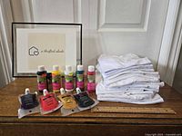 Overview photo showing stack of 16 white children's t-shirts and 10 fabric paint containers on a wooden table with rulers for scale.