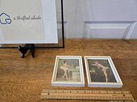 Set of two white framed ballerina prints positioned side by side on a wooden surface with a ruler for scale.