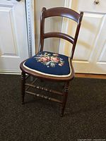 Front and angle view of wooden accent chair with hand-stitched needlepoint floral seat upholstery on dark blue background, showing carved backrest and turned legs.