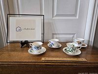 Four teacups and matching saucers displayed on wooden surface with measuring ruler, showing floral designs in blue hydrangea, purple wildflowers, lily of the valley, and pink-orange roses. Tea sets in good condition with no visible chips or cracks.