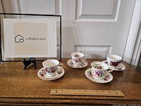 Image of 4 floral patterned teacups each with matching saucers arranged in a row with a ruler for scale.