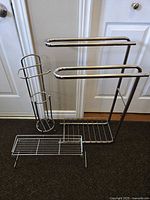 Three chrome bathroom caddies photographed side-by-side against a white door background on a carpeted floor, showing a toilet paper holder, towel rack with two bars, and a wire bath caddy shelf