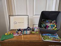 Three Lego Friends small building sets displayed on table along with instruction booklet and loose parts in fabric bin.