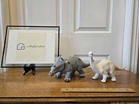 Two ceramic dinosaur figurines shown side by side with a ruler for scale, one in grey glaze and the other white and unfinished.