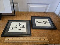 Two small framed Inuit art prints on a wooden surface with a ruler showing the scale. One print shows five flying geese and the other a dog sled scene.
