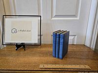 Three blue cloth hardcover books by Thornton W. Burgess standing upright on a wooden surface next to a framed logo for a thrifted abode.