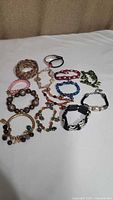 Overview photo showing 12 assorted costume jewelry bracelets in various colors and styles including faux pearls, braided mesh, heavy beads, and garden-themed charms.