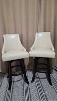 Two cream genuine leather swivel bar stools with wooden legs shown from front.