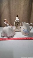 Three ceramic items: figurine, clock, and pitcher displayed with a ruler for size reference