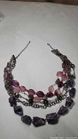 Photo of a multi-strand necklace on a white surface, showing purple rough stones, pink and purple beads, and silver-tone chain links.