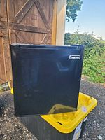Closed black Magic Chef mini refrigerator shown on yellow bin outdoors.