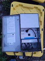 Outdoor lighting transformer with the metal cover open showing wiring terminals, power outlet, and control switches inside the enclosure.