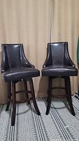 Pair of two espresso colored leather swivel bar chairs in front of curtain, showing overall shape and design.