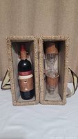 Open rattan carrying case with one wine bottle on left side held by two leather straps, and two stacked wine glasses on right side with leather holders securing them.