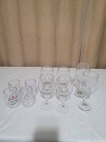 Six clear heavy glass stemmed wine glasses with classic shape in front with four plastic stemless Christmas-themed glasses behind.