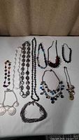 Photo of 11 assorted costume jewelry necklaces on a white surface, various beaded, strand, and multi-strand styles visible.