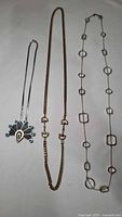 Three necklaces laid out showing large turquoise pendant, gold plated horse bit chain, and silver-tone geometric link chain