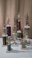 Photo showing nine vintage sports trophies with gold-tone athlete figurines on colorful striped marble bases against brown curtain background.