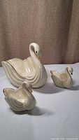 Photo shows three ivory ceramic swan candy dishes of different sizes with gold accents on rim and beak, arranged on white surface.