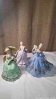 Group photo of three ceramic figurines in green, blue, and lavender dresses.
