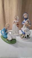 Three ceramic figurines: girl with geese, girl with lamb, woman with basket and lamb together; handpainted details
