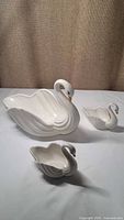 Three white ceramic swan candy dishes of varying sizes displayed on a white surface with a textured brown background.