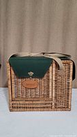 Wicker picnic and bar basket with green top flap and beige cloth strap against neutral background.