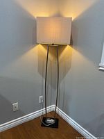 Beige floor lamp illuminated in a corner, showing warm light emission from a rectangular fabric shade and slender metallic post above a black square base.