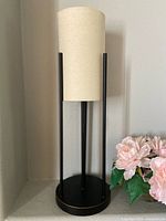 Round lamp with cylindrical beige shade supported by black metal rods on a round base, photographed with floral decor.