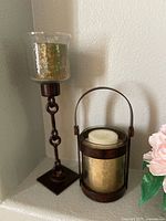 Tall metal candle holder with glass hurricane cover and smaller lantern-style candle holder with handle, both containing candles, situated on a corner shelf.