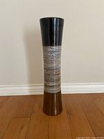Full view of a tall ceramic floor vase with black top and brown textured midsection placed on wooden floor against white wall.