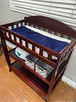 Full view of wood baby changing table in espresso cherry finish with navy blue changing pad, safety rails, and storage shelves containing baby wipes and baskets.