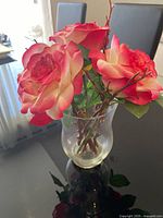Glass vase with artificial silk red and cream roses and leaves, with twine around the vase neck, on a dark reflective surface in front of chair and window.