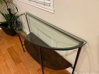 Clear top-down and front-angle view of glass top accent table showing the half-moon shape glass top, black metal frame, and wooden bottom shelf in dark brown finish.