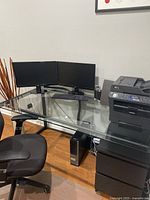 Photo showing L-shaped glass top desk with triple monitor setup, ergonomic office chair, and black steel frame supporting toughened glass desktop.