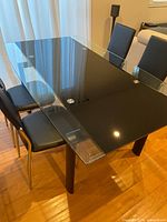 View of glass top dining table with black metal legs and four black upholstered chairs with chrome legs surrounding the table on wood flooring near window with sheer curtains.