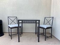 Front view of outdoor metal framed square table and two matching chairs with cushions.