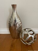 Front view of silver bud vase next to hollow silver decorative sphere on wooden floor by white wall