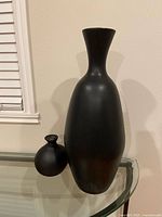 Two black ceramic vases, one tall and slender with flared top, and one small round, displayed on glass surface against white wall.
