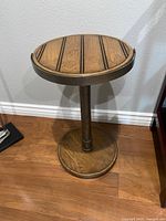 Side view of the light brown wooden round side table showing the single center leg and circular base on wood floor against a gray wall.
