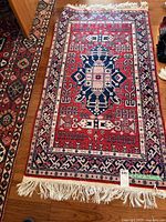 Full view of the small rectangular rug laid out on a wood floor, showing the geometric patterned design in red, navy blue, and cream with fringed edges.