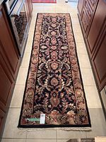 Full view of rectangular ornate floral area rug on kitchen floor, black base with red and beige pattern, fringed edges visible.