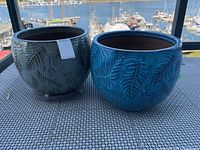 Two ceramic pots placed side-by-side on a table with a harbor background; shows blue pot on right and gray pot on left.
