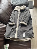 Gray long coat shown flat on a brown couch, with visible cream-colored faux shearling trim on collar, cuffs, front edge, and hem.