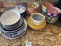 A collection of colourful bowls and plates on a kitchen counter. Includes patterned bowls with bright floral and geometric designs, cream and yellow bowls, and blue and white floral plates.
