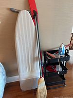 Photo showing white ironing board, broom leaning against wall, and black three-tier trolley cart with iron on top shelf.