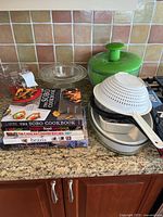 Photo showing stacked cookbooks, mixing bowls in metal and plastic, a white colander, and a green lidded bowl on countertop.