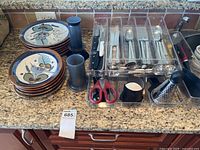 Sets of ceramic dinner and side plates with blue, green, brown floral leaf pattern, alongside a clear plastic tray with stored cutlery and kitchen tools on countertop.