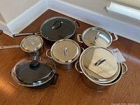 Full set of pots and pans arranged on wooden floor, showing all items with lids included.